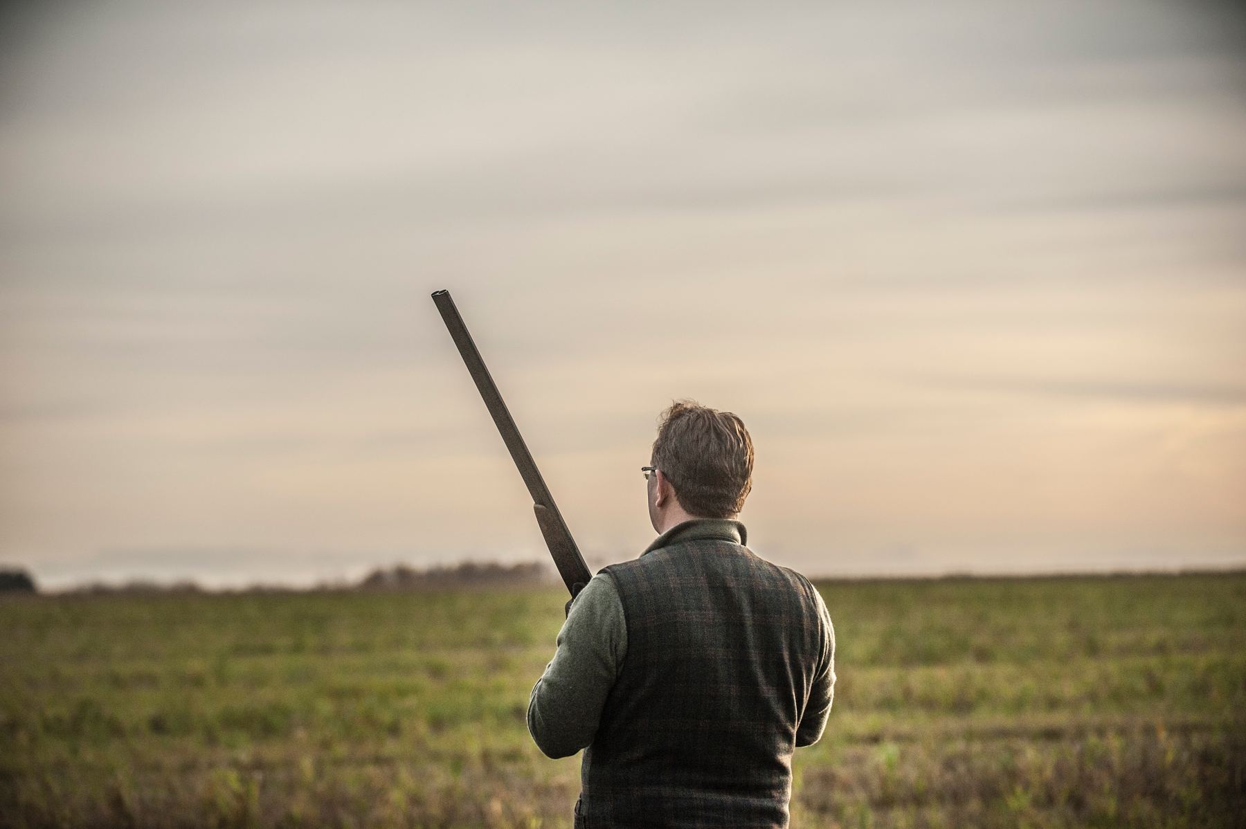 A gun with his shotgun over his shoulder looking out across open Suffolk farmland at dusk